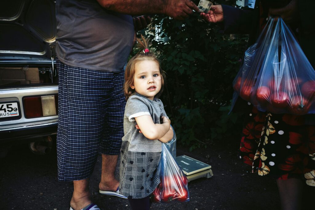 Una niña encantadora sostiene una bolsa de productos en un mercado al aire libre, irradiando inocencia.