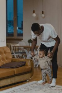 A father assists his toddler in walking inside a cozy living room.